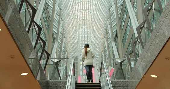  Descrição da imagem: podemos observar na imagem uma das saídas do sistema PATH cidade subterrânea Toronto, onde uma escada rolante central leva uma mulher para um dos túneis acima da terra. O túnel possui o teto tomado por vidros, o que proporciona uma belíssima iluminação natural.